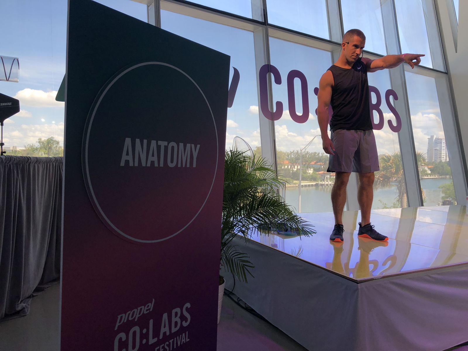 Grant Weeditz pointing from a glass stage beside an Anatomy sign, with a view of the bay behind him at Faena, Miami Beach